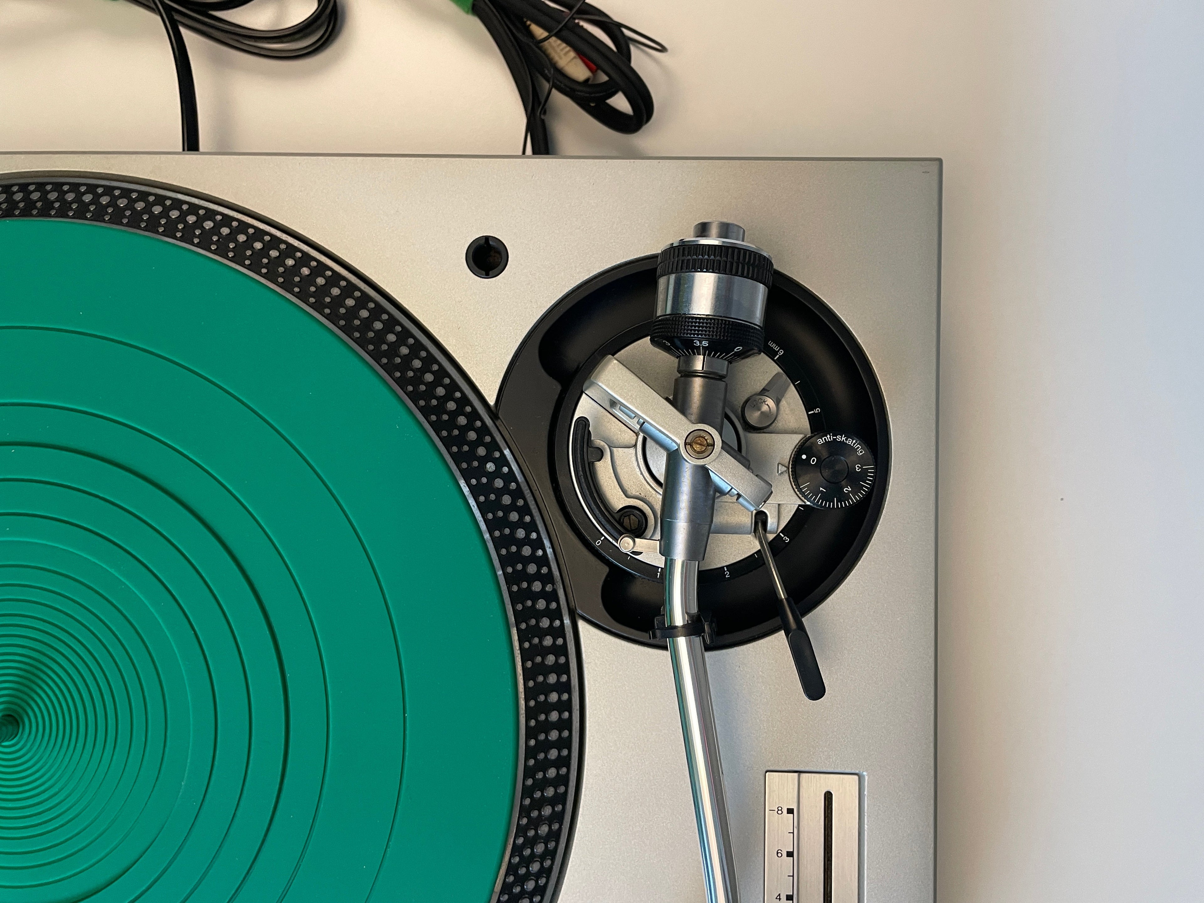 Close-up of a silver turntable with a bright green platter and black outer ring, showing part of the tonearm and control dial against a white background.
