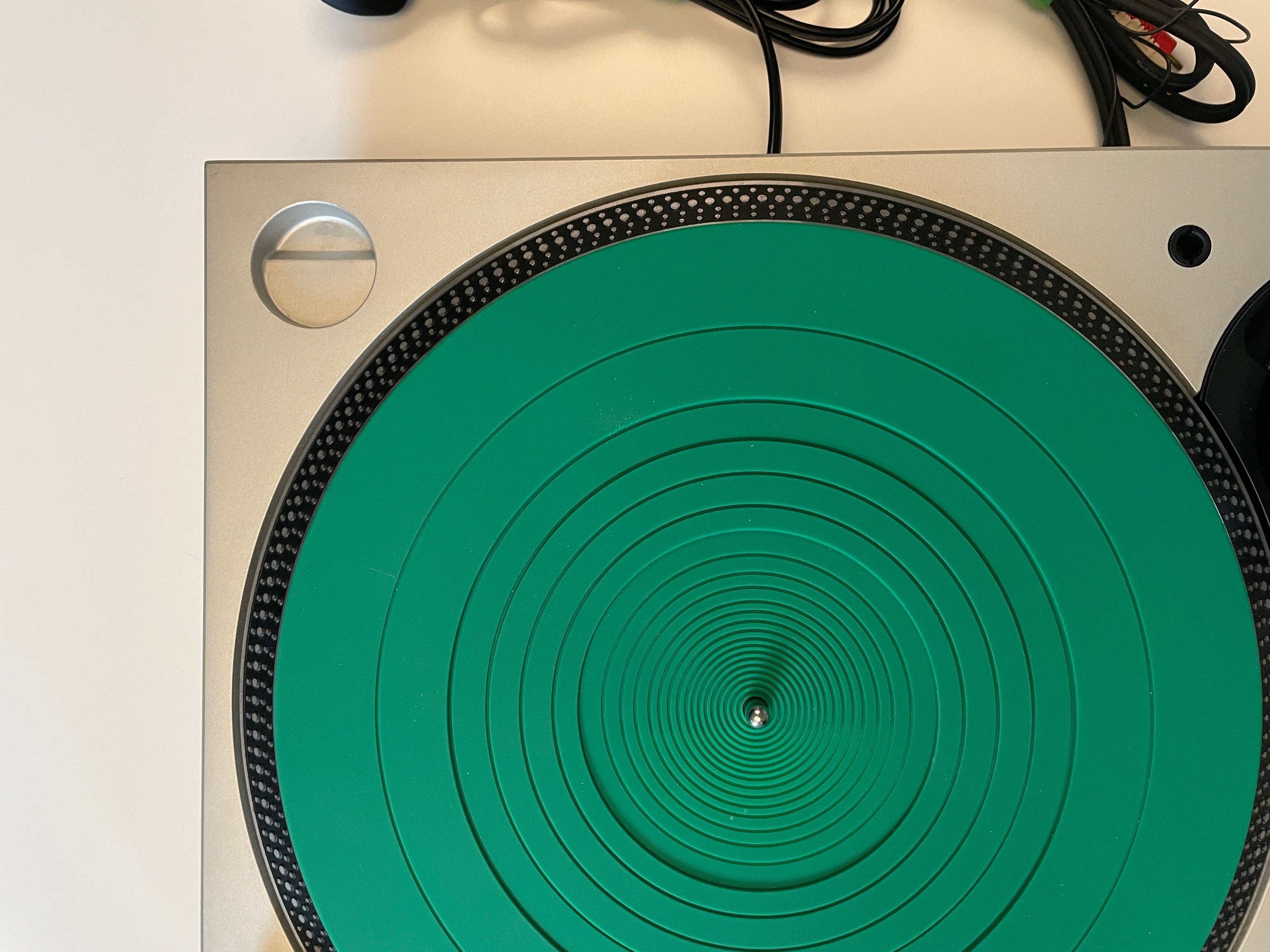 Top-down view of a turntable with a bright green plinth and a silver metallic base, photographed on a light neutral background with some cables visible at the bottom.