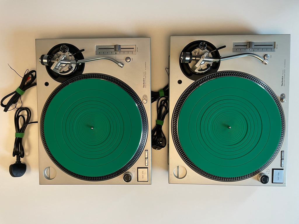 Two silver turntables with green rubber mats, shown side by side on a plain light background. Coiled black cables are visible on both units.