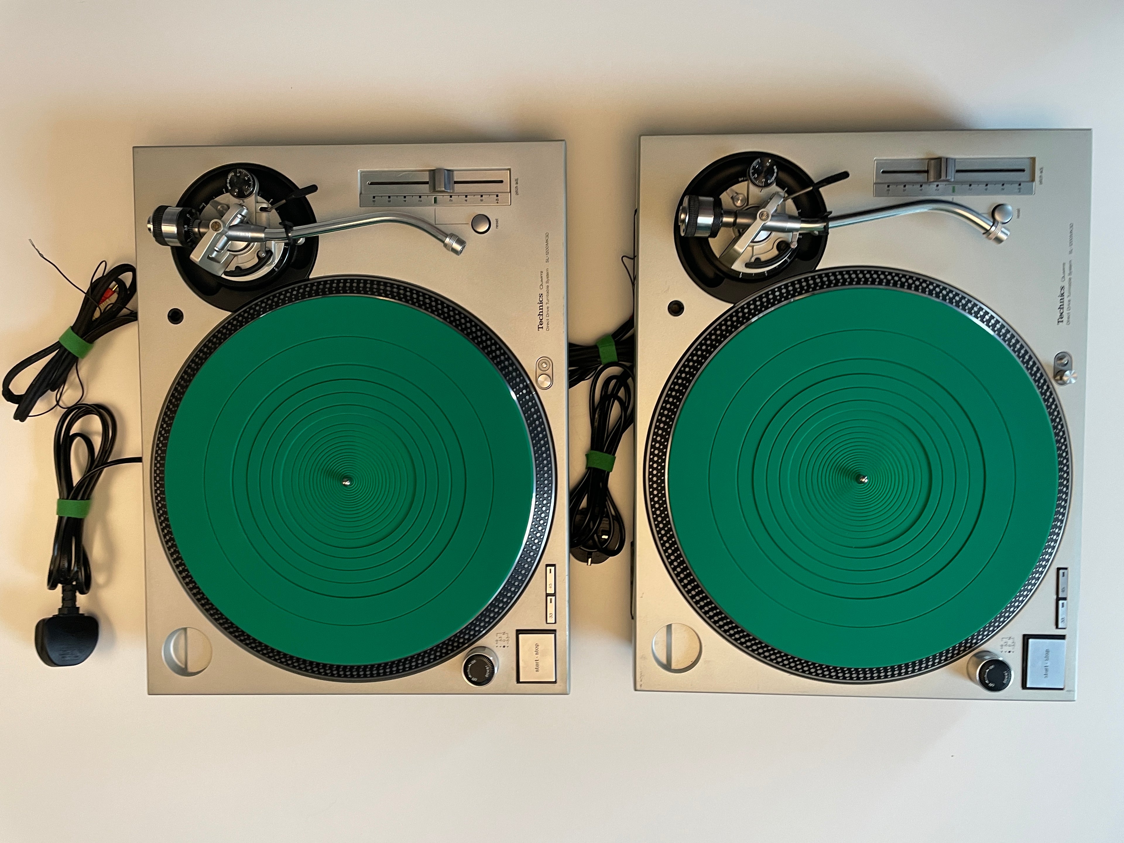 Two silver turntables with green rubber mats, shown side by side on a plain light background. Coiled black cables are visible on both units.