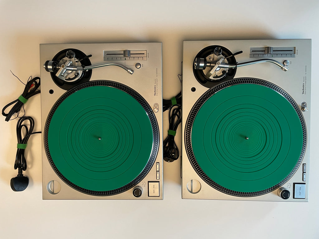 Two silver turntable-style record players with large green platters, shown side by side on a plain light background with attached power cords.