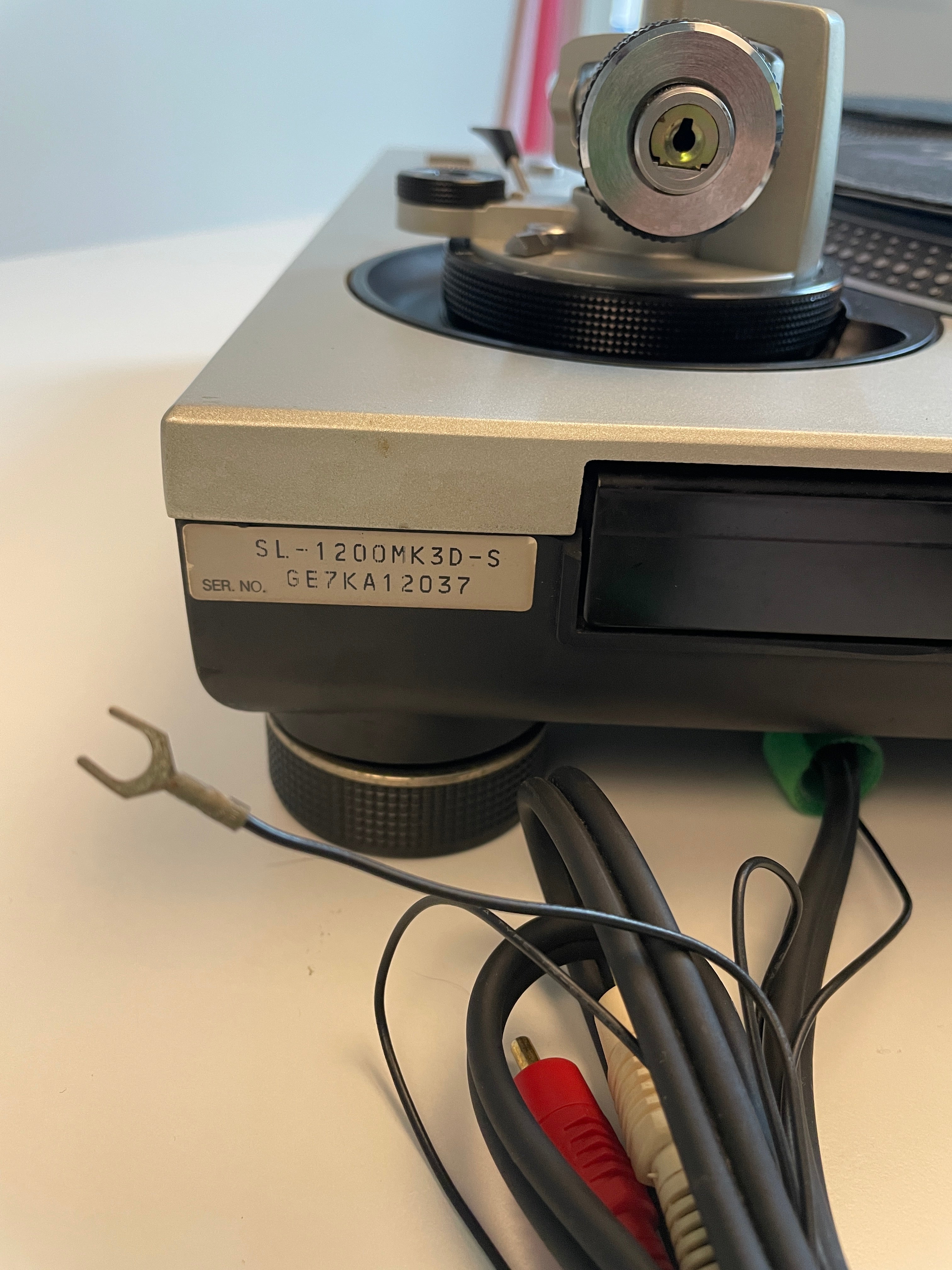 A close-up of a vintage audio equipment chassis with a silver and black metal body, a model label reading SL-1200MK3D-S, and attached audio cables with red, white, and black connectors on a white surface.