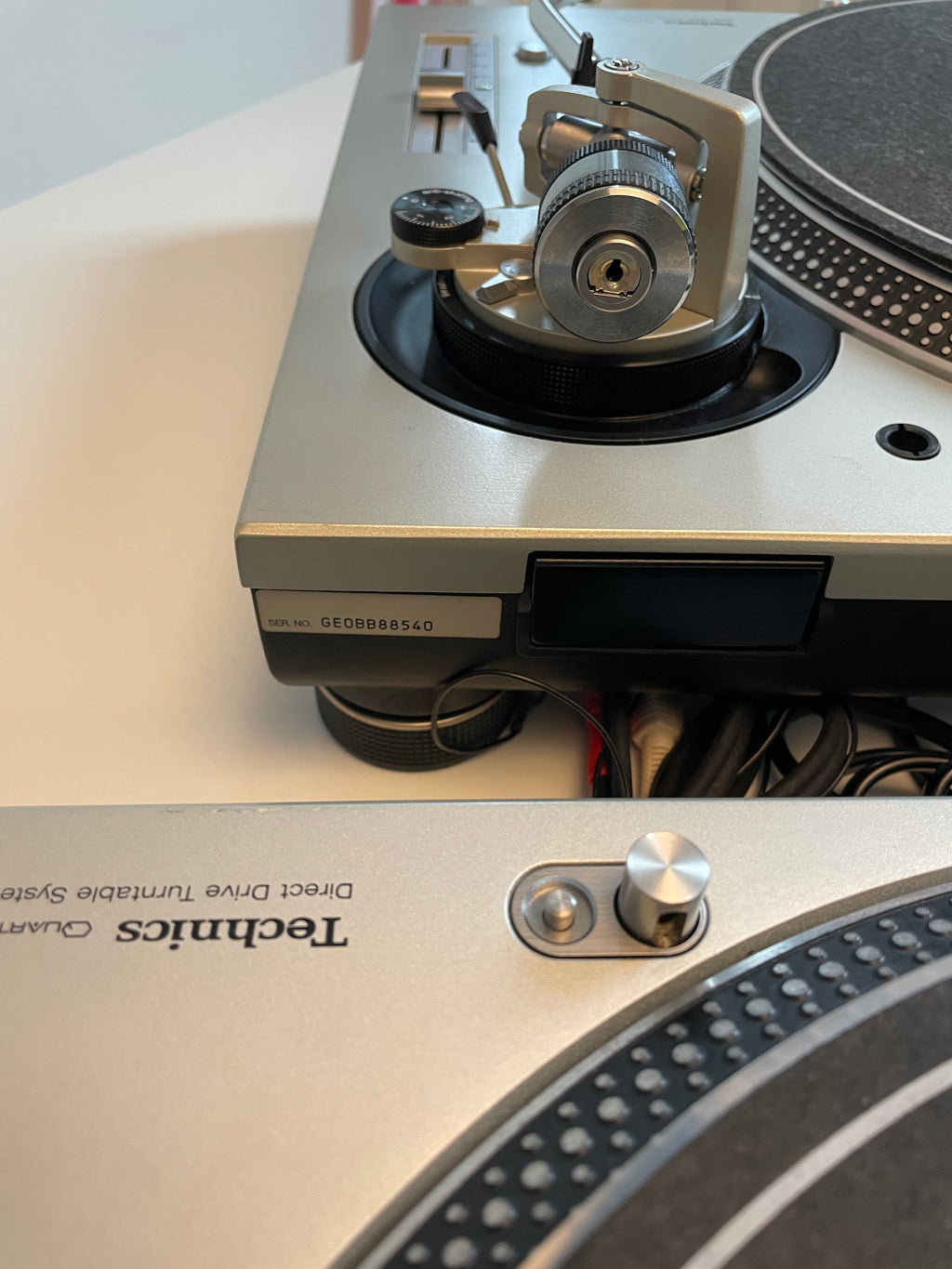 Close-up of a Technics turntable showing the silver and black plinth, tonearm, cartridge, and part of the platter, resting on a white surface.
