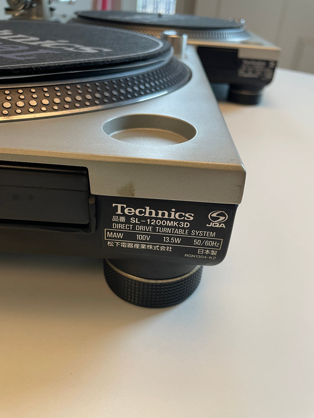 Close-up of a Technics SL-1200MK3D direct drive turntable system on a white surface, showing the side label and part of the platter and tonearm.