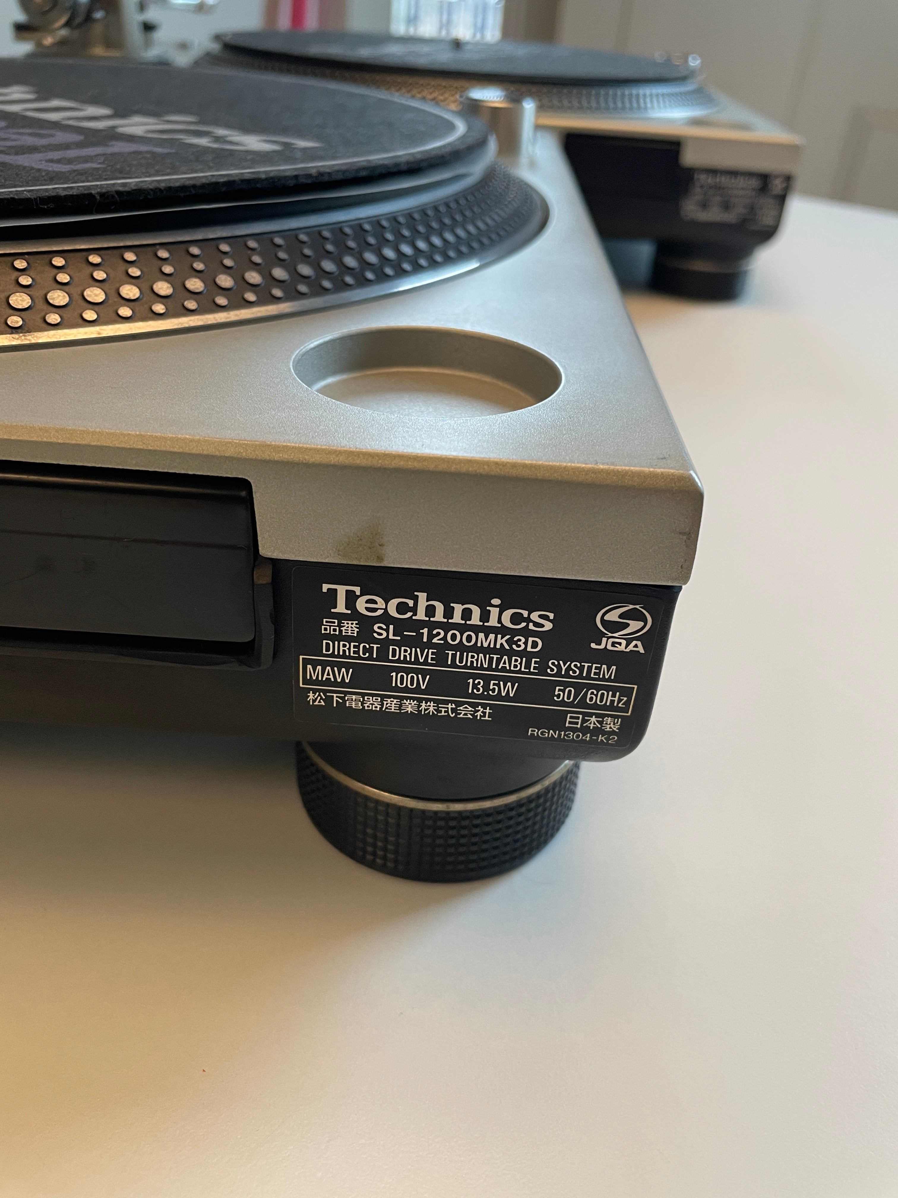 Close-up of a Technics SL-1200MK3D direct drive turntable system on a white surface, showing the side label and part of the platter and tonearm.