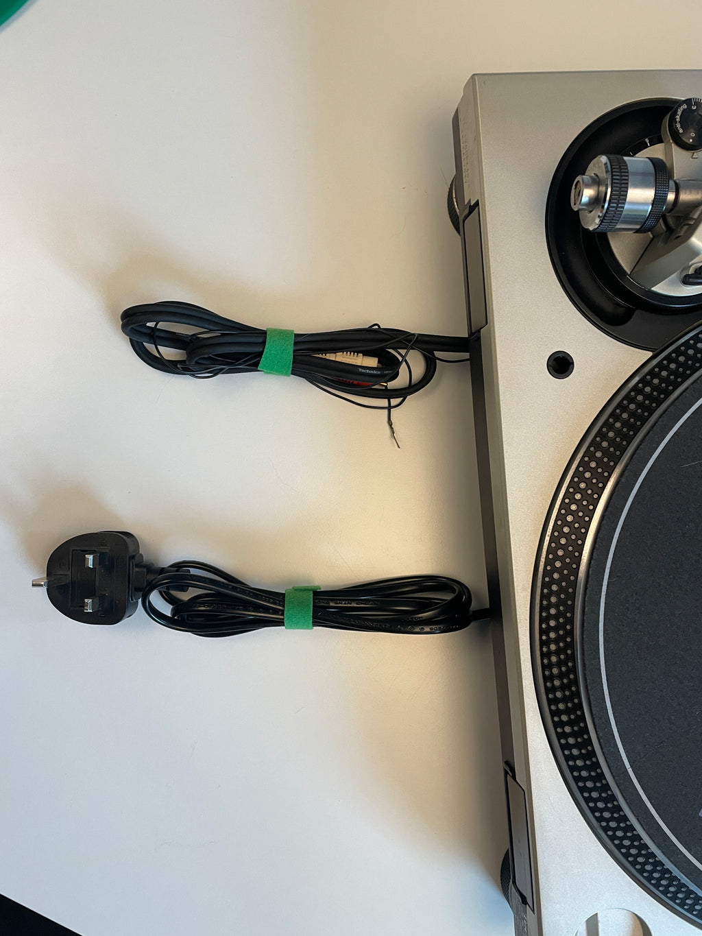 A close-up of a silver and black turntable showing its power cord and audio cable, which are bundled with green zip ties and have a UK two-pin plug.
