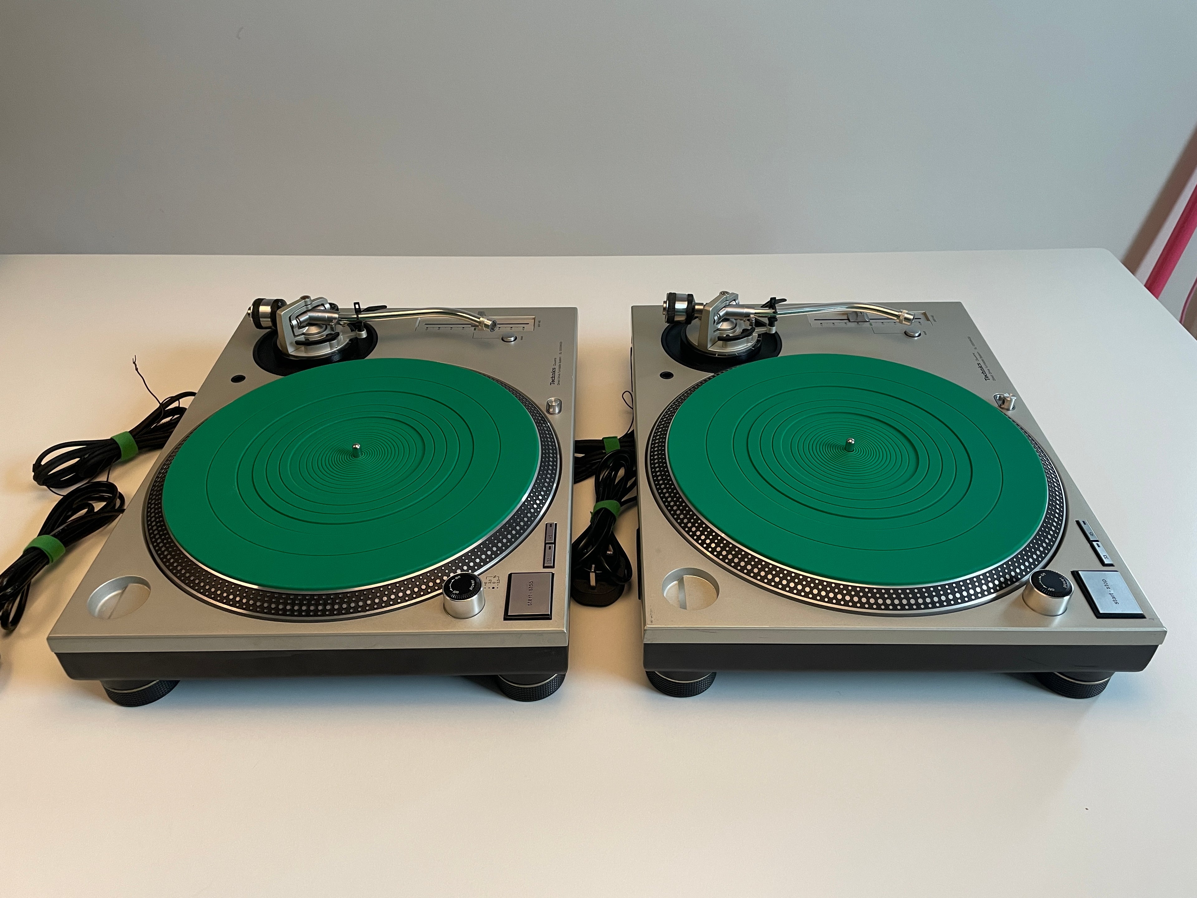 Two silver turntable players with bright green plinth covers, shown side by side on a white tabletop against a light gray wall.