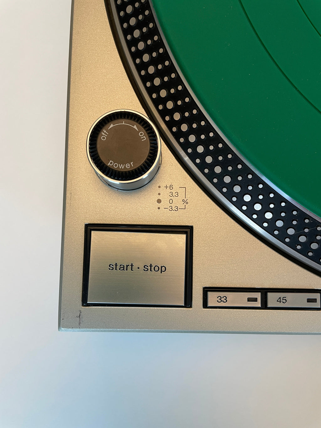Close-up of a turntable control panel showing a silver start/stop button, power dial, and a green vinyl record partially visible against a plain light background.
