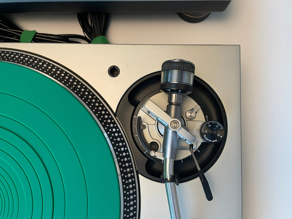Close-up of a turntable showing the silver and black tonearm, plinth, and a bright green vinyl record surface.
