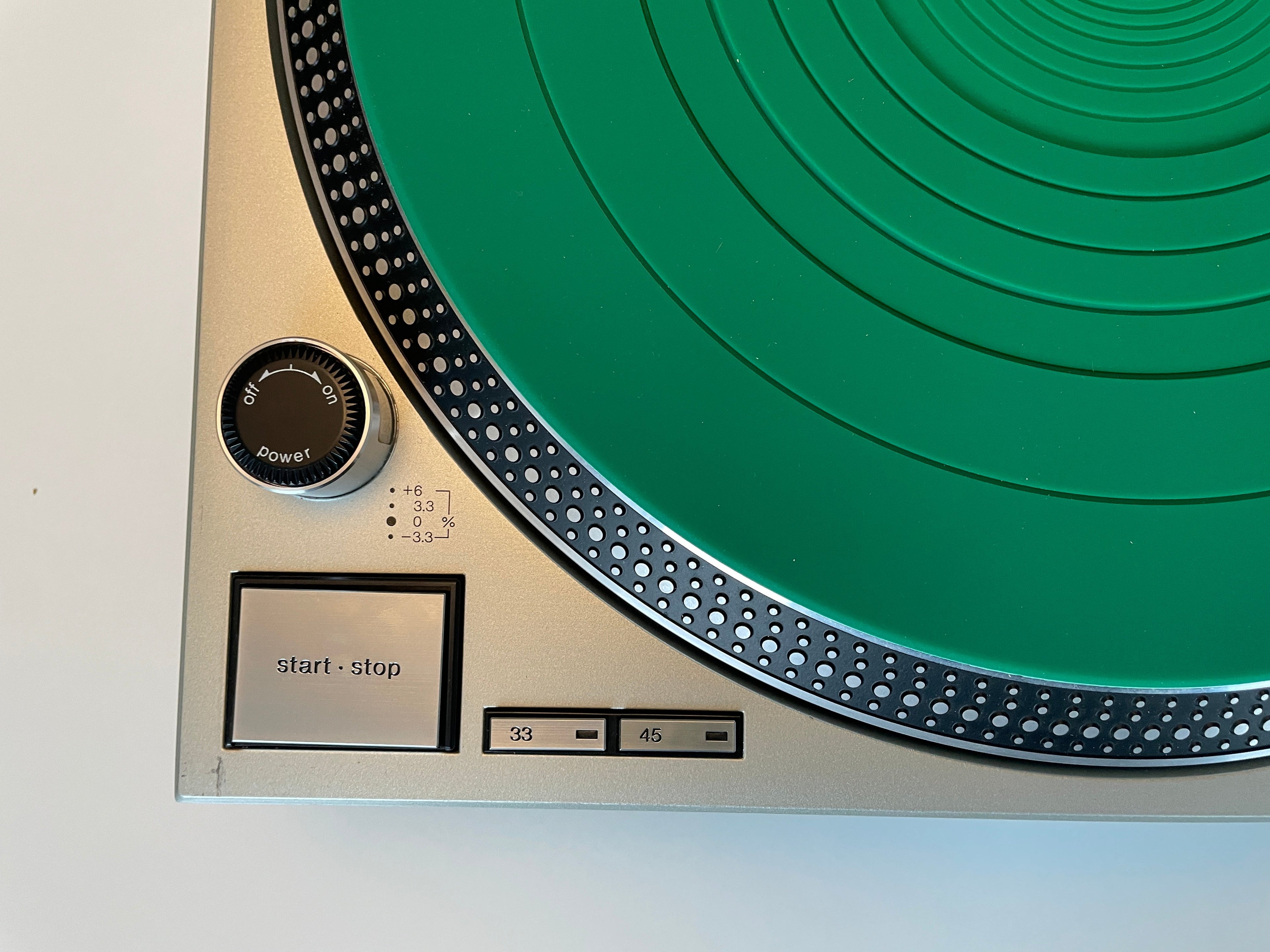 Close-up of a turntable control panel with a large green vinyl record partially visible, a metallic beige body, black buttons, and a dial on a plain light background.