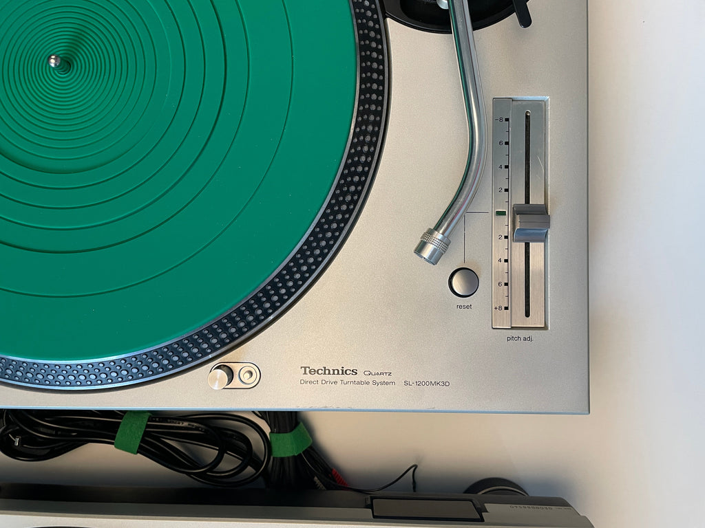 A close-up of a Technics SL-1200 turntable with a silver metal chassis, featuring a bright green vinyl record on the platter and a silver tonearm.