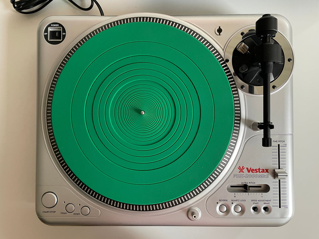 A silver Vestax turntable with a prominent green circular plinth, photographed from directly above on a light surface.