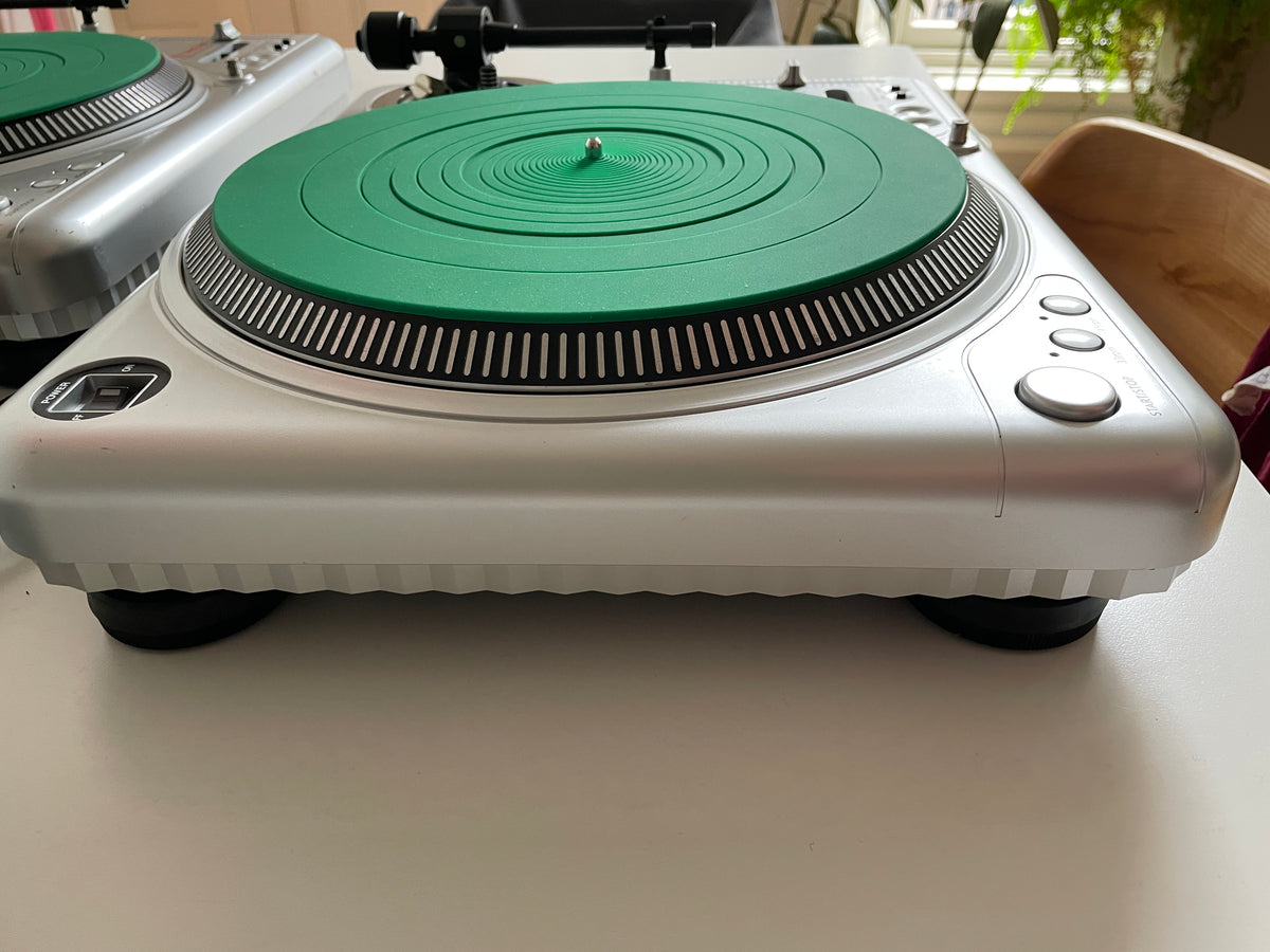 A silver turntable with a distinctive green circular plinth and matching green protective cover, shown on a white tabletop with another turntable partially visible in the background.