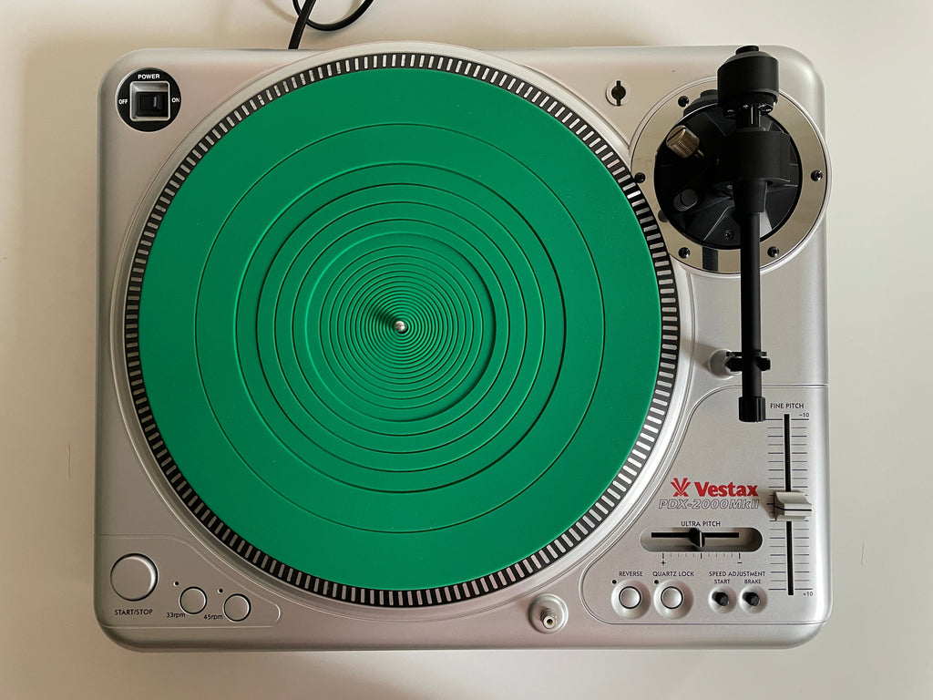 A silver Vestax turntable with a distinctive green circular vinyl record skin, photographed from directly above on a white surface.
