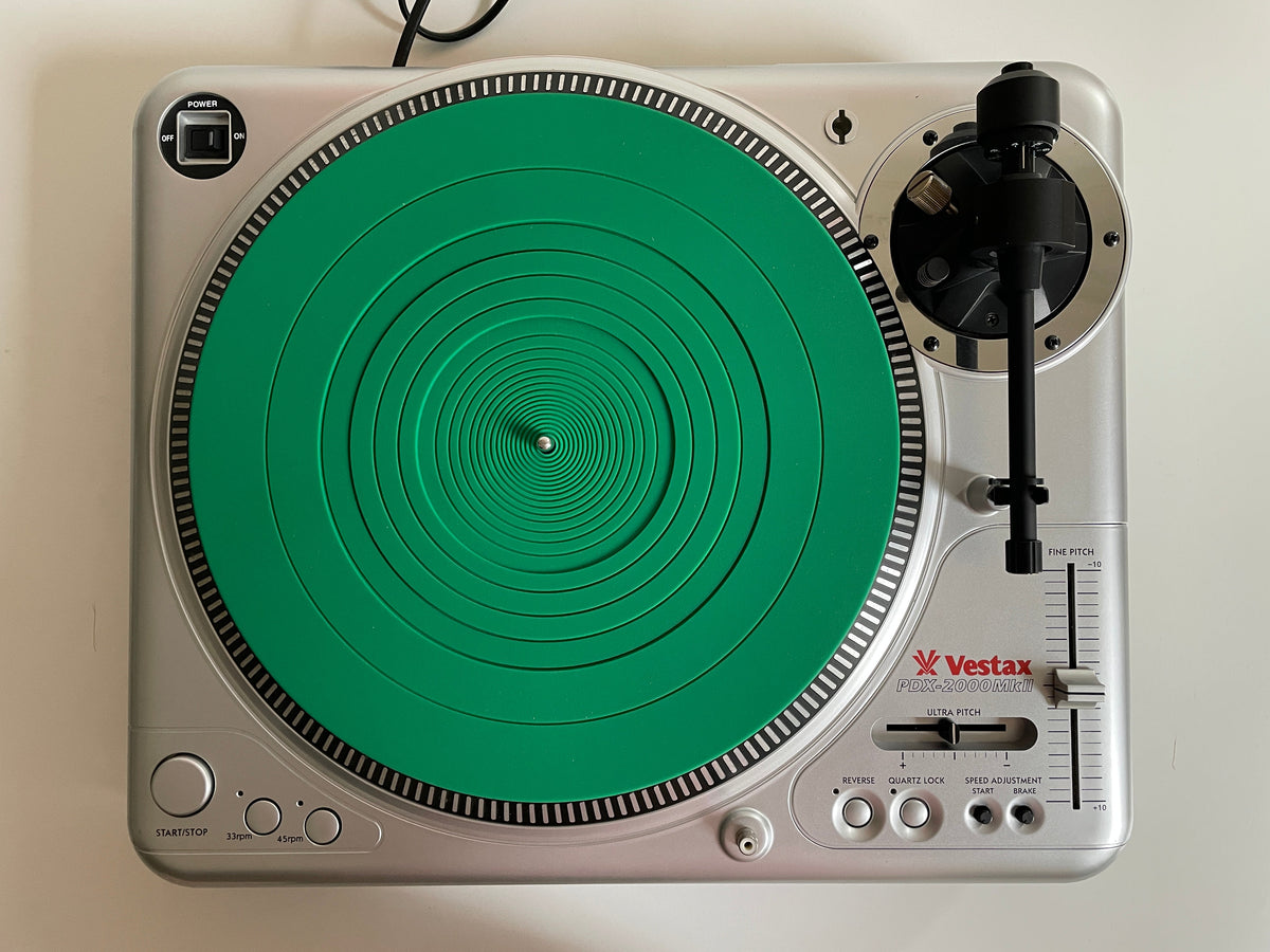A silver Vestax turntable with a distinctive green circular vinyl record skin, photographed from directly above on a white surface.