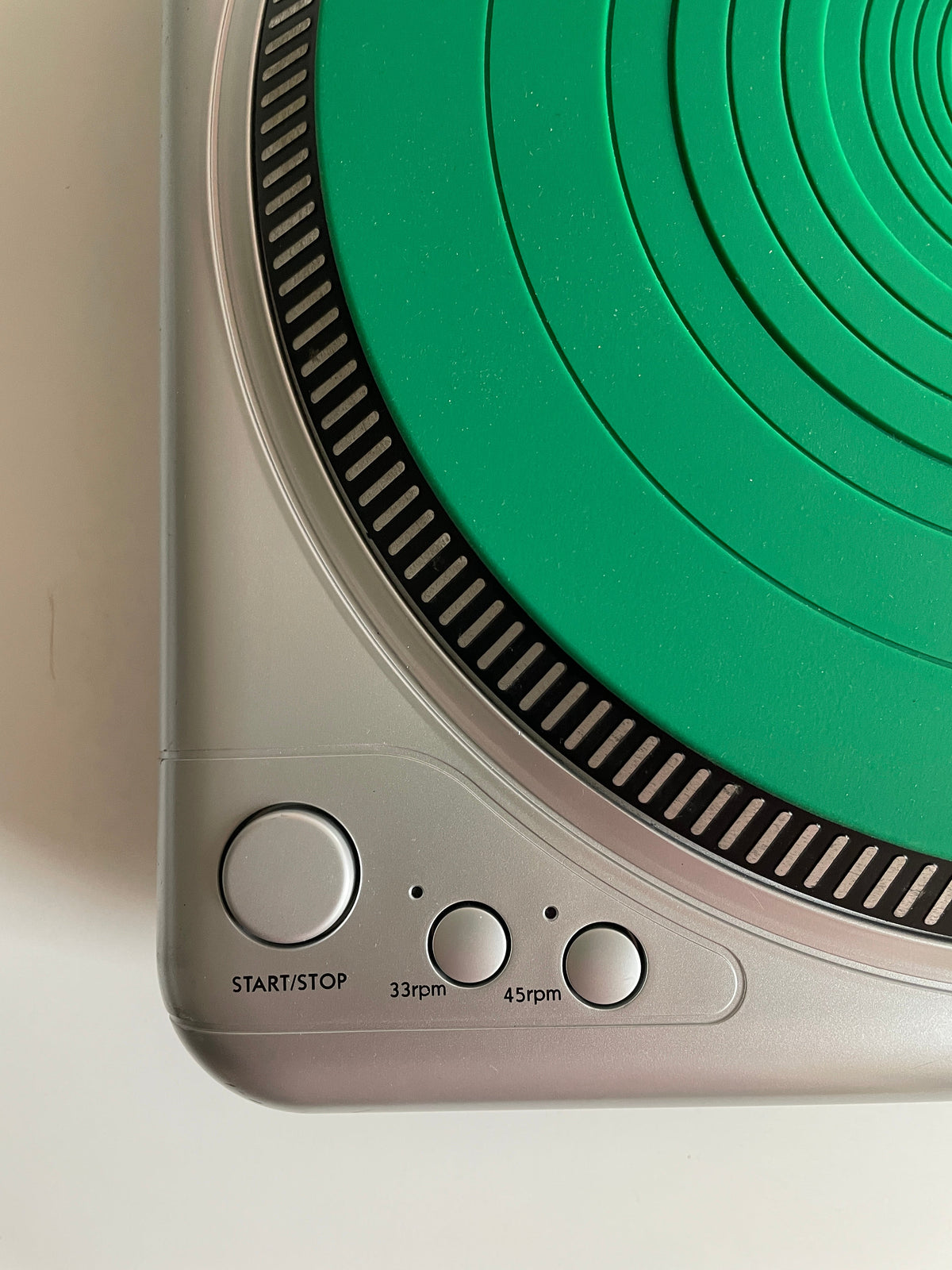 Close-up of a silver and green portable speaker or electronic device, showing the circular green driver area, black striped ring, and side buttons labeled 45rpm, 33rpm, and START/STOP.