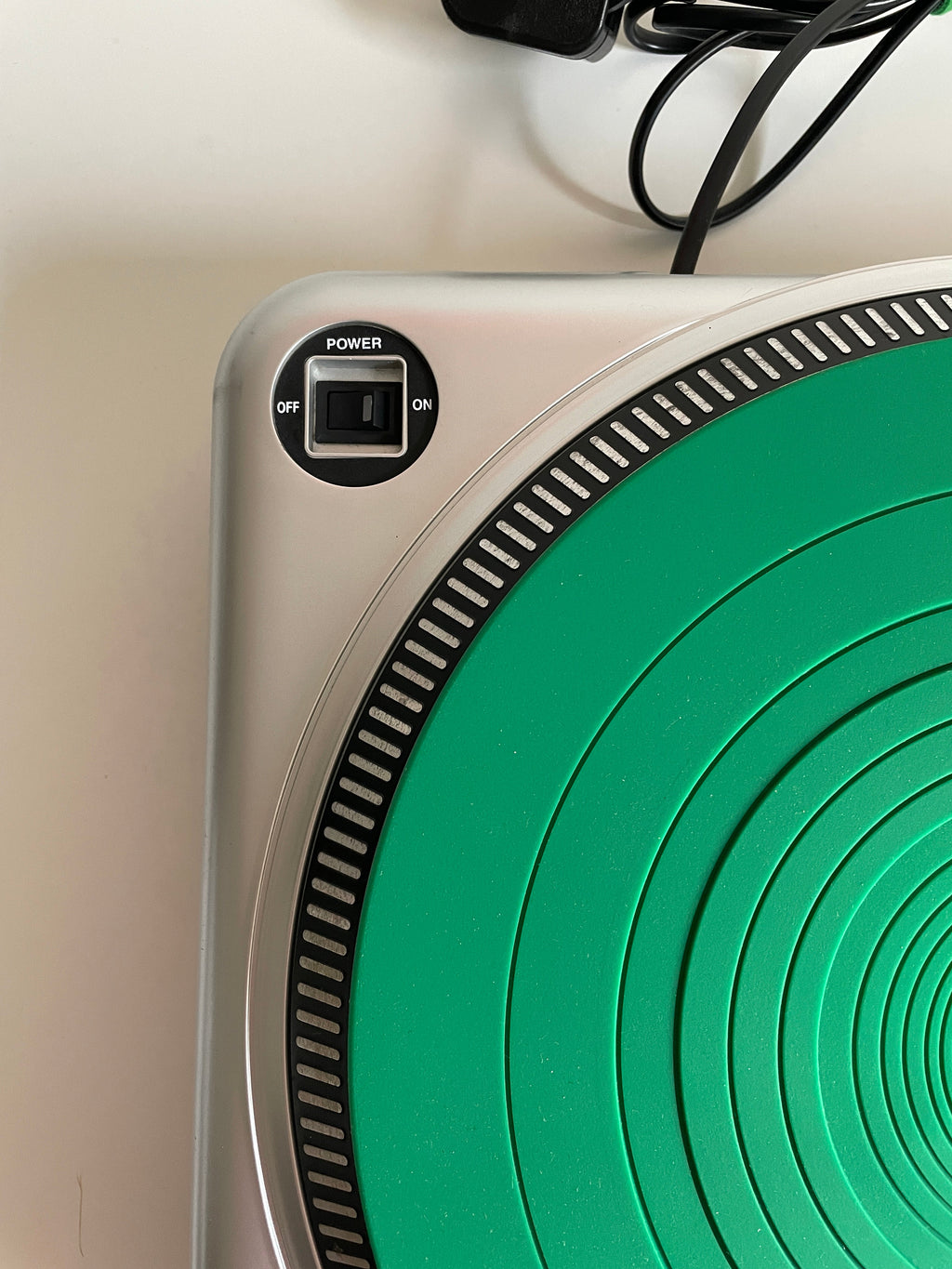 Close-up of a portable electric heater or hot plate with a silver base, a green circular heating plate, a power button, and a black power cord on a light surface.