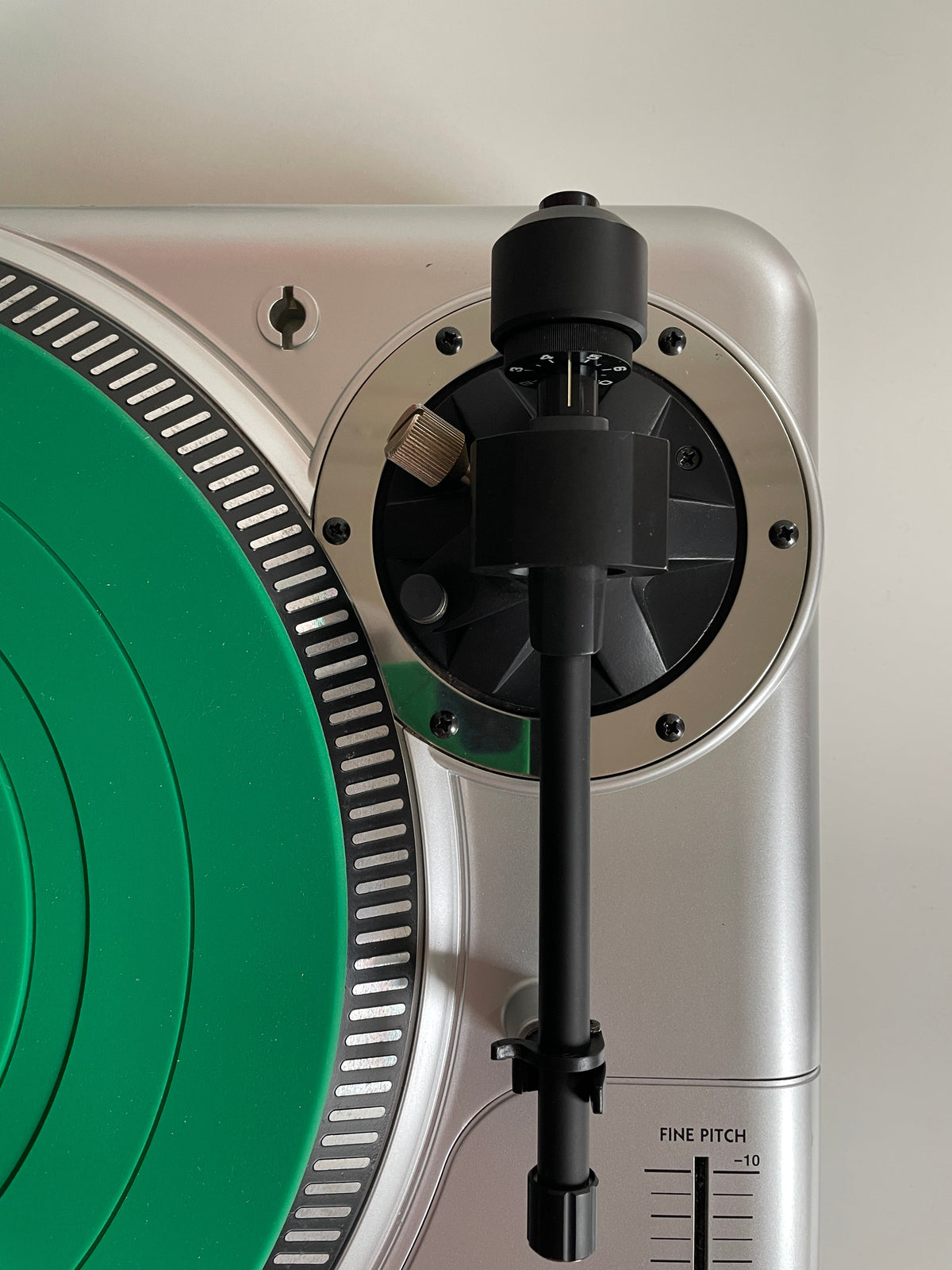 Close-up of a silver turntable showing the tonearm, inner tonearm mechanism, and a green vinyl record partially visible.
