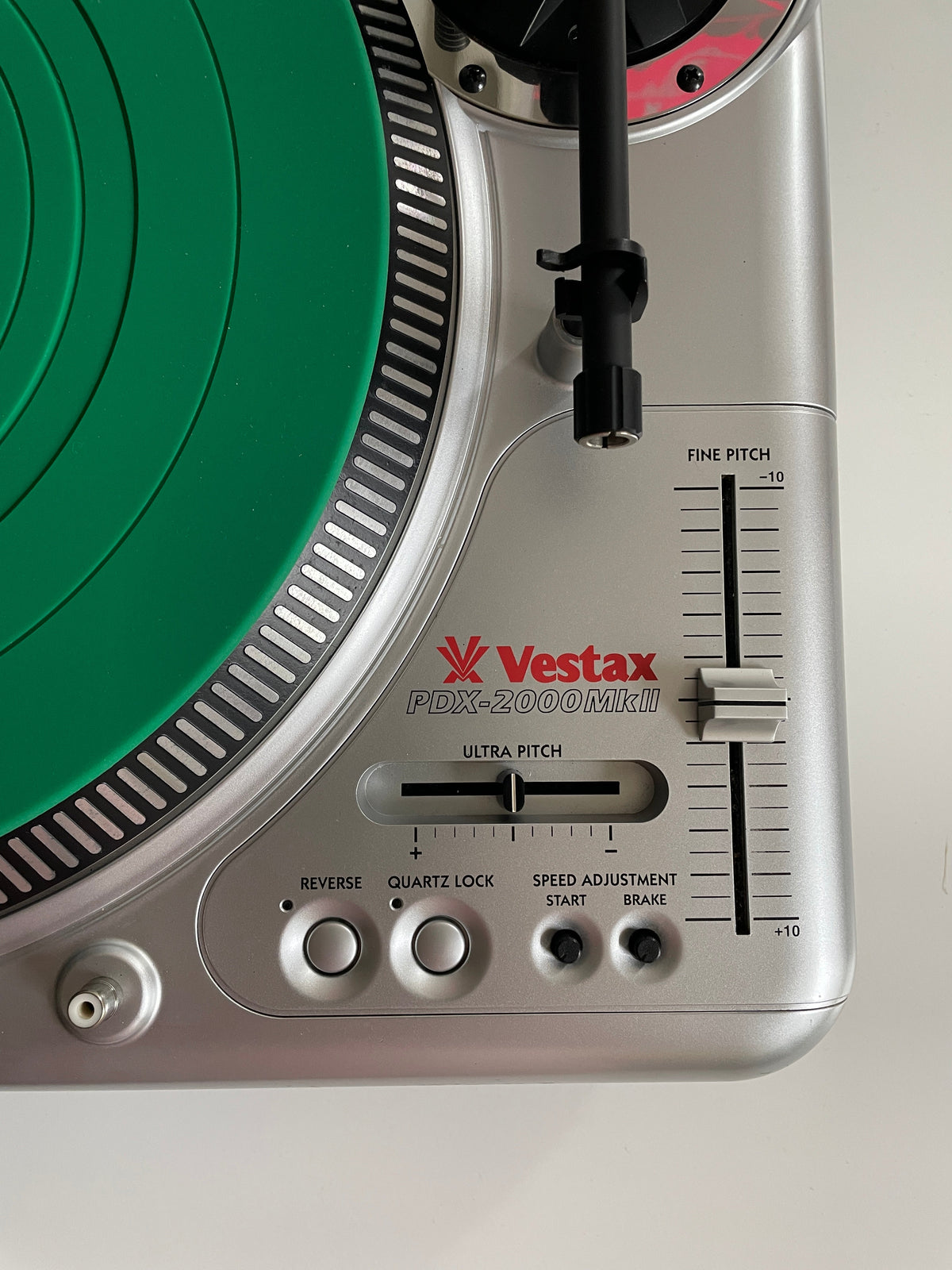 Close-up of a silver Vestax PDX 2000 MK2 turntable showing the tonearm, control buttons, and a green vinyl record on a plain light background.