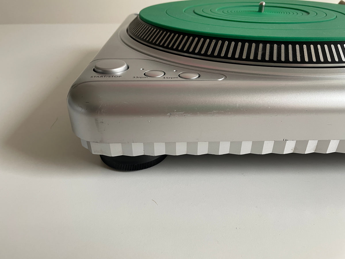 A close-up of a silver turntable or record player base with a green vinyl record on the platter, photographed on a plain white surface.
