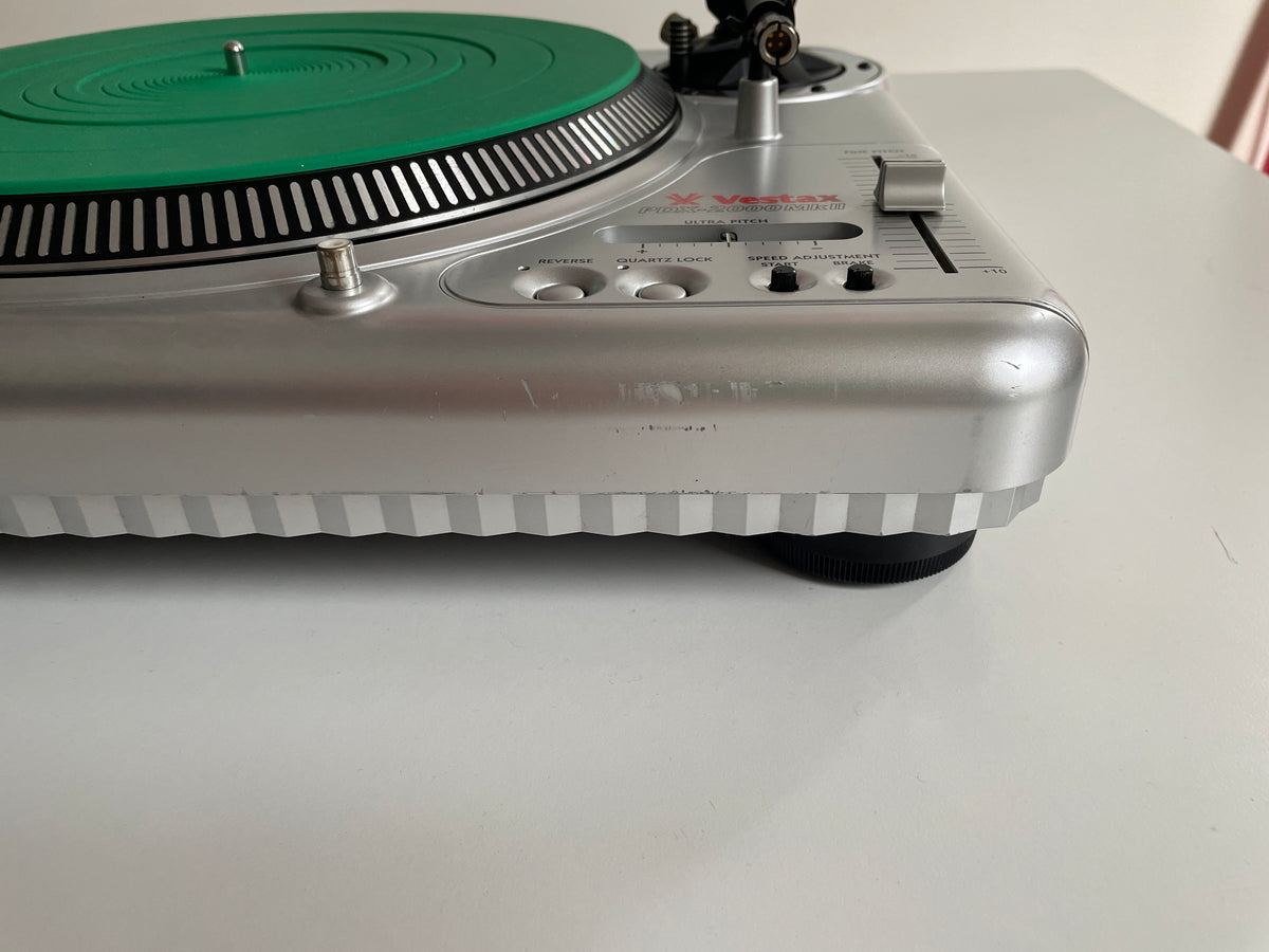 A close-up side view of a vintage Vestax turntable, showing its silver chassis, green plinth, and black tonearm on a white tabletop.