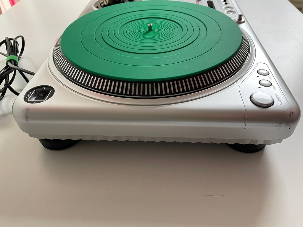 A silver and green turntable or CD player photographed from above on a white surface, with a power cord visible on the left.