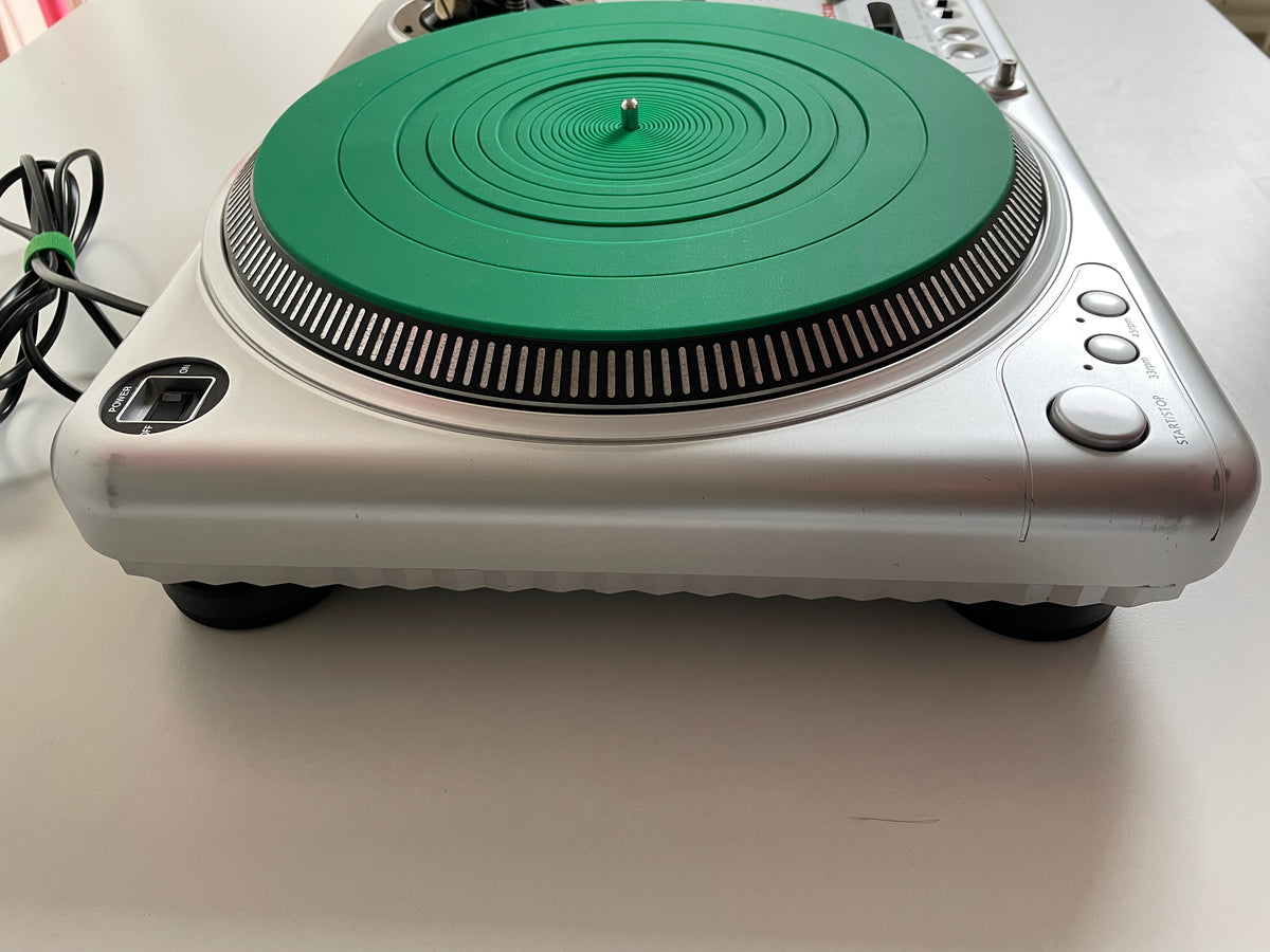 A silver and green turntable or CD player photographed from above on a white surface, with a power cord visible on the left.