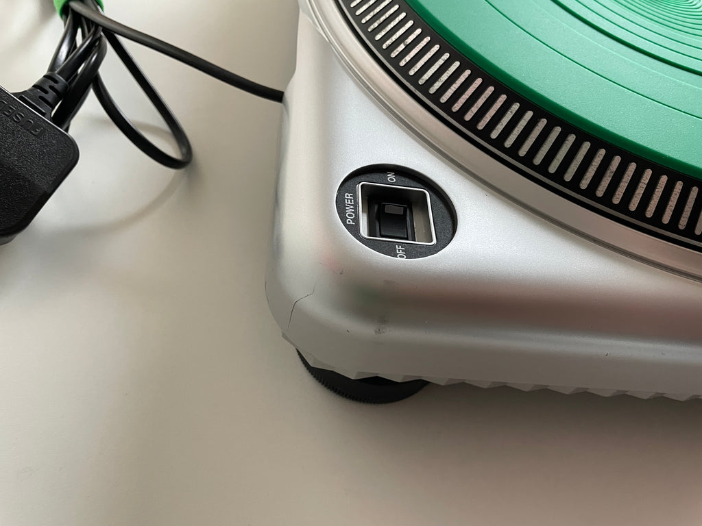 Close-up of a silver and green portable turntable showing the power button and power cord on a light surface.