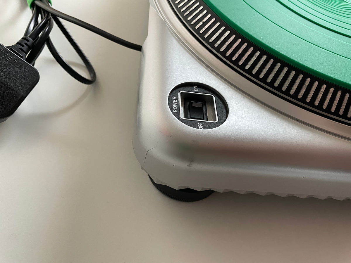 Close-up of a silver and green portable turntable showing the power button and power cord on a light surface.