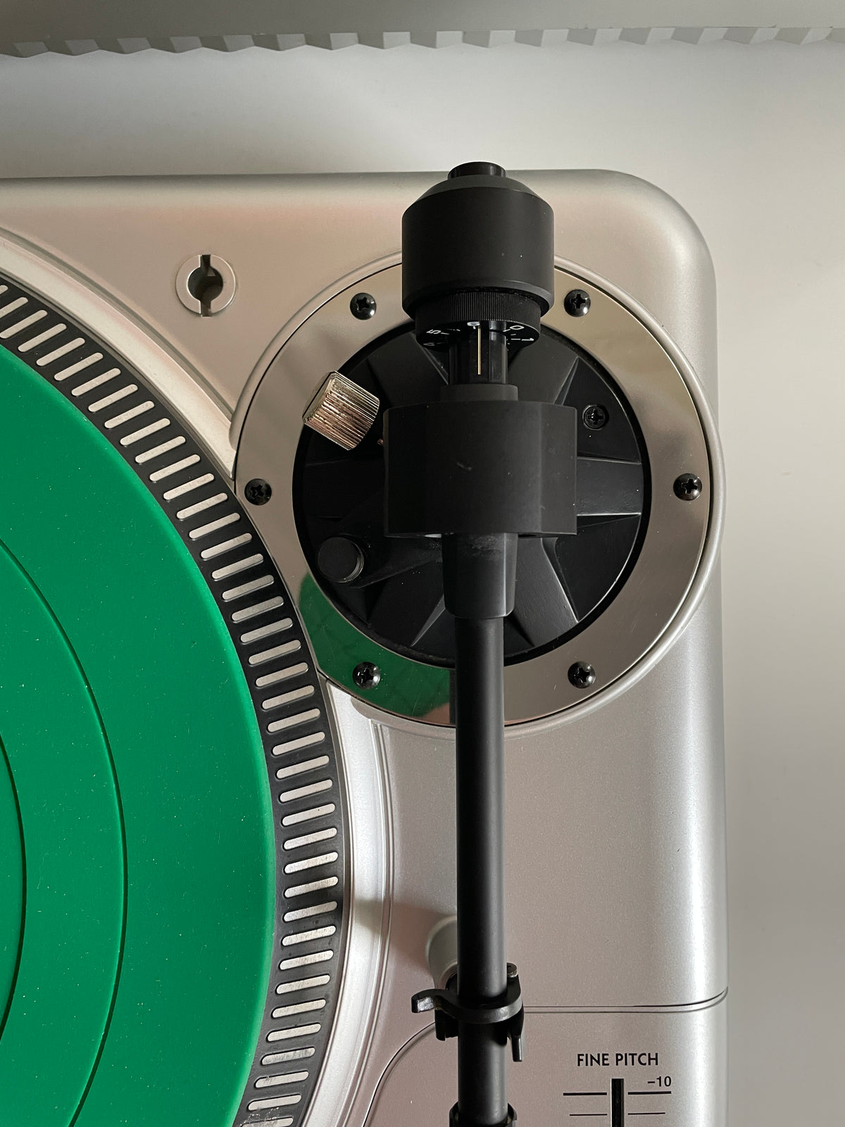 Close-up of a silver and black turntable component with a green circular platter edge visible, showing the tonearm and internal mechanism.