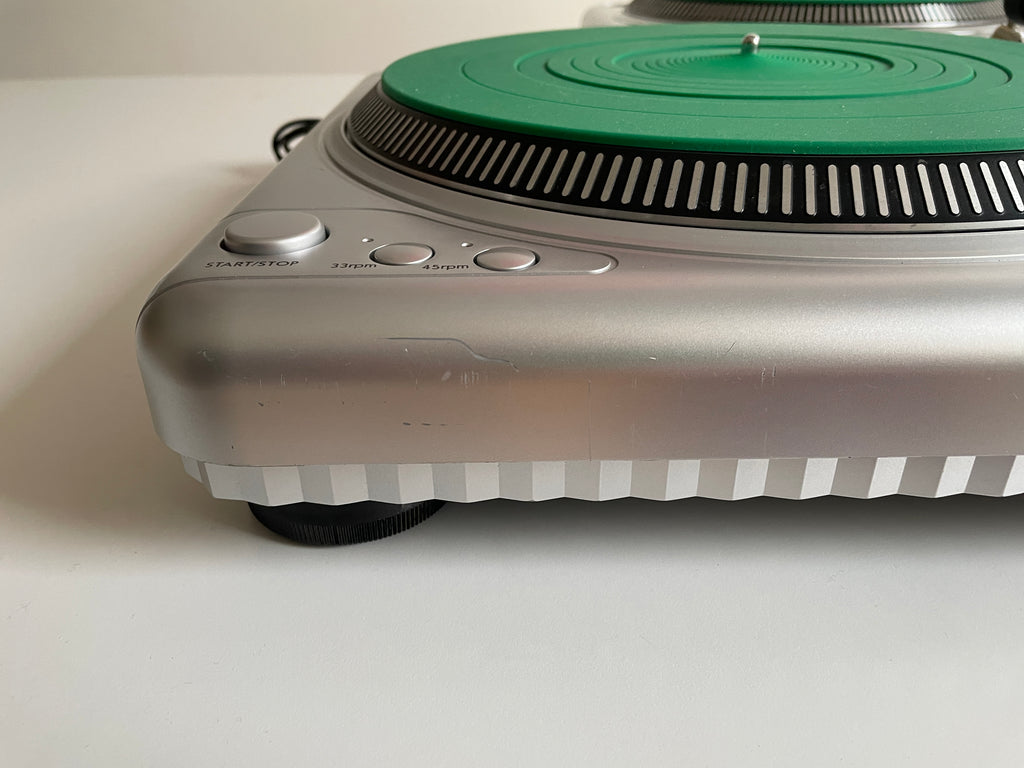 Close-up of a silver turntable showing the side control buttons, a green platter, and part of the white outer flange on a white surface.