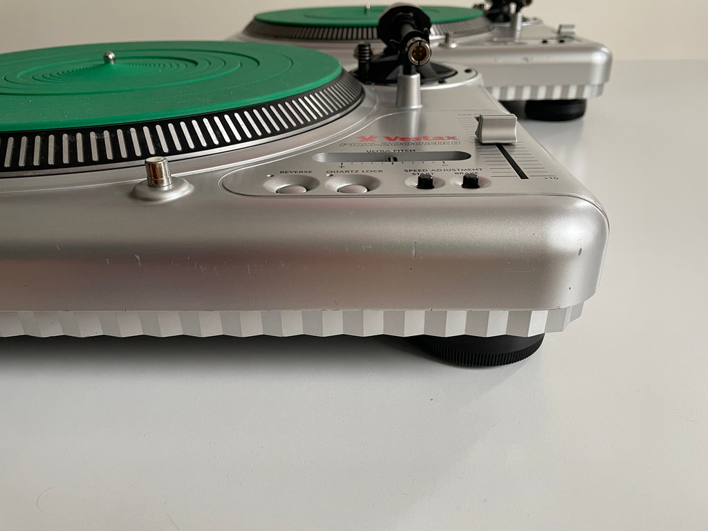 A close-up of a silver turntable with a green platter, with two more turntables blurred in the background on a white surface.