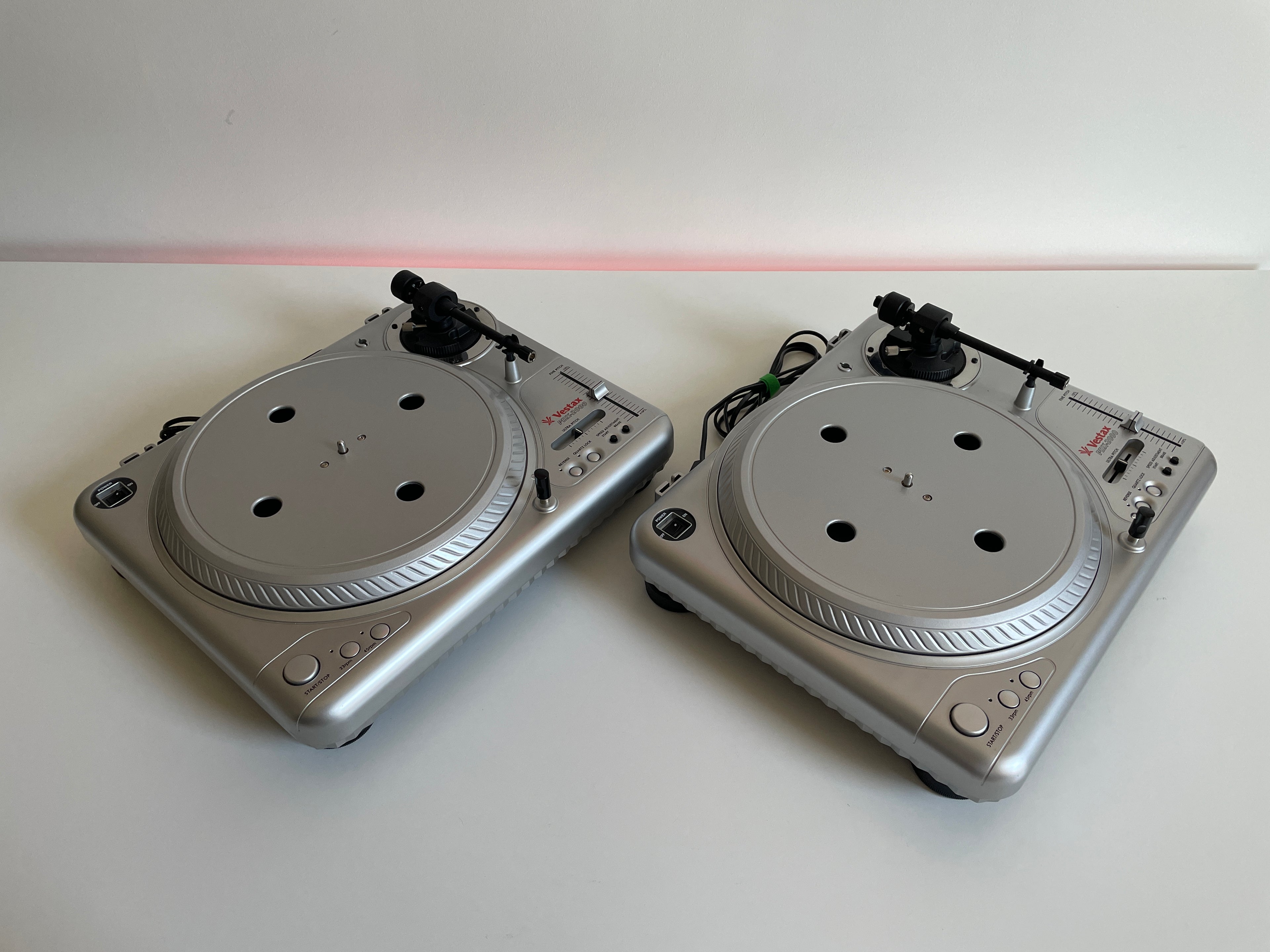 Two silver DJ turntables on a white surface with a red light in the background.